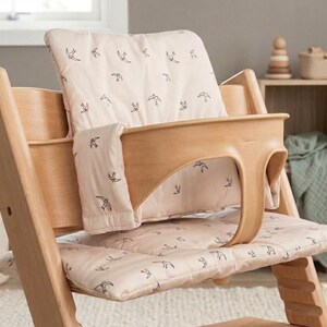 May include: A light wood high chair featuring a two-piece padded cushion set in a soft beige color. The cushions are decorated with a repeating pattern of small, dark-colored bird silhouettes. The high chair includes a curved wooden safety rail.