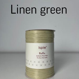 Puede incluir: Un carrete de rafia de color verde claro etiquetado como Linen green. La etiqueta de papel blanco presenta la marca Ispie, el texto Raffia, Authentic Plant Fiber, The Most Silky Raffia y dos filas de pequeñas cruces grises en el frente.