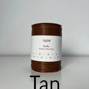 Puede incluir: Una bobina de rafia de color tostado de la marca Ispie, etiquetada como fibra vegetal auténtica. La bobina cilíndrica está envuelta en una banda de papel blanco con el nombre de la marca, el tipo de producto y un patrón decorativo de pequeñas cruces grises.