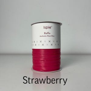 Puede incluir: Un carrete de rafia de la marca Ispie en un color rojo fresa vibrante. La etiqueta blanca presenta el texto Ispie, Raffia, Authentic Plant Fiber y un patrón de pequeños símbolos x grises. La palabra Strawberry está impresa en negro debajo.