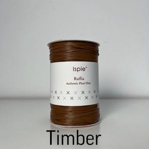 Puede incluir: Un carrete de rafia, fibra vegetal auténtica, de color marrón con una etiqueta blanca que presenta el texto Ispie, Raffia, Authentic Plant Fiber y dos filas de pequeños símbolos x. La palabra Timber está impresa en negro debajo.