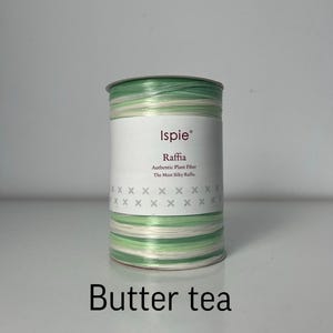 Puede incluir: Un carrete cilíndrico de fibra vegetal de rafia de la marca Ispie en una paleta de colores variados de verde claro, verde oscuro y blanco. La etiqueta de papel blanco presenta el texto Ispie, Raffia, Authentic Plant Fiber, The Most Silky Raffia y Butter tea.