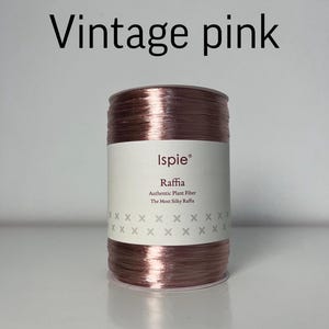 Puede incluir: Un carrete de fibra vegetal de rafia en color rosa vintage con una etiqueta blanca que presenta el texto Ispie, Raffia, Authentic Plant Fiber, The Most Silky Raffia y un patrón de pequeñas cruces grises. El carrete es cilíndrico y metálico.