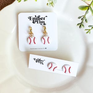 Könnte beinhalten: Weiße Baseball-Ohrringe und Ohrstecker mit roten Nahtdetails. Die Ohrringe befinden sich auf einer weißen Karte mit dem Text "Ember Elm Handmade in Kansas City". Die Ohrringe befinden sich auf einem weißen Teller.