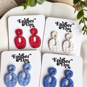 Op de afbeelding: Vier paar hangende oorbellen op witte displaykaarten met het label Ember & Elm. De oorbellen hebben getextureerde, ovale hangers in rood, wit, lichtblauw en middenblauw. Elk paar bestaat uit een klein rond bovenstuk en een groter ovaal.