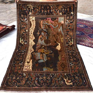 Peut inclure: Un grand tapis rectangulaire brun foncé avec un motif complexe. Le panneau central présente une scène détaillée avec des personnages et des animaux dans des tons de marron, de bleu et de rose. La bordure est ornée de motifs floraux et d'oiseaux. D'autres tapis sont visibles en arrière-plan.