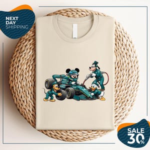 May include: A beige t-shirt featuring a graphic of Mickey Mouse in a green Formula 1 race car, with Goofy, Pluto, and Donald Duck dressed in matching green pit crew uniforms. The car displays the number 14 and Aston Martin branding.