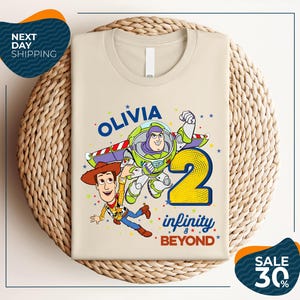 A t-shirt featuring a character from the Toy Story franchise, specifically Buzz Lightyear, along with the text "Olivia 2 Infinity Beyond". The t-shirt is displayed on a woven basket, and there are two sale signs indicating a 30% discount.