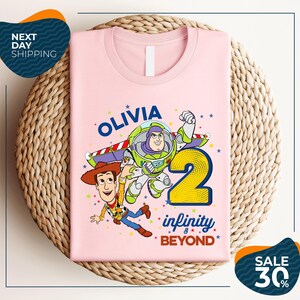 A pink t-shirt with a cartoon image of Buzz Lightyear and Woody from Toy Story, along with the text "Olivia 2 Infinity Beyond".