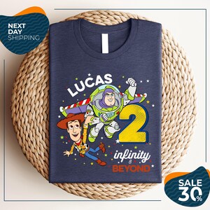 A blue t-shirt with a graphic design featuring characters from the Toy Story franchise, specifically Buzz Lightyear and Woody, along with the text "Lucas 2" and " Infinity & Beyond".