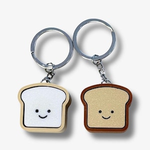 Op de afbeelding: Twee sleutelhangers met bedels in de vorm van een sneetje brood. De ene bedel is wit met een lichtbruine rand, de andere is lichtbruin met een donkerbruine rand. Beide hebben een simpel zwart gezichtje met twee stippen en een mondje.