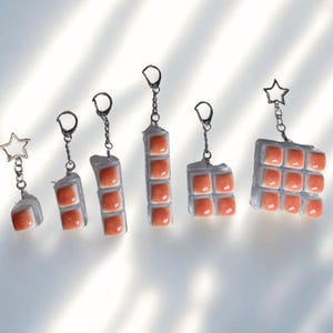 Könnte beinhalten: Eine Sammlung einzigartiger Schlüsselanhänger mit transparenten, rechteckigen Harzblöcken mit orangefarbenen quadratischen Akzenten. Jeder Schlüsselanhänger hat eine silberne Kette und einen silbernen Stern- oder runden Verschluss.