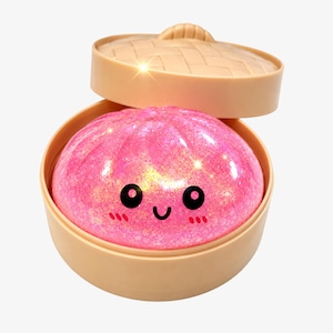 May include: A bright pink, glittery, squishy dumpling toy with a smiling face, black eyes, and red cheeks, resting inside a light tan, round, plastic bamboo-style steamer basket with a matching lid tilted open at the top.