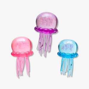 May include: Three translucent jellyfish figurines in purple, pink, and blue. Each jellyfish features a rounded, glitter-filled bell top with a scalloped edge and multiple thin, dangling tentacles extending downward from the base of the bell.