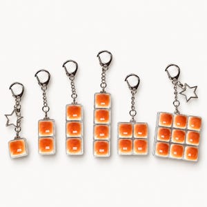 May include: A collection of six silver-toned keychains featuring orange, square, resin-like tiles arranged in various grid patterns. Some keychains include a small silver star charm attached to the chain, and all have a metal lobster clasp.