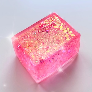 Könnte beinhalten: Ein rechteckiger Block aus durchscheinendem rosa Harz, der eine dichte Konzentration von eingebetteten goldenen und rosa Glitzerpartikeln im gesamten Material aufweist. Das Objekt hat eine glatte, glänzende Oberfläche mit scharfen Kanten.
