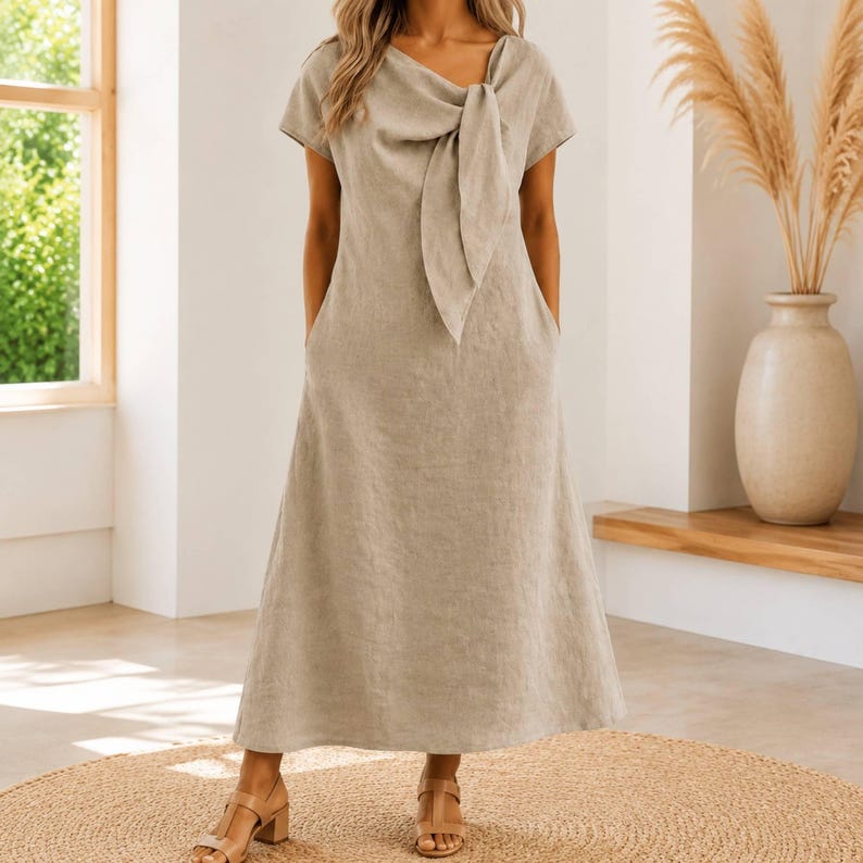 May include: A beige midi-length dress with short sleeves, a round neckline, and a prominent fabric tie detail at the center of the chest. The dress features a straight, flowing silhouette that extends to the mid-calf and includes side pockets.