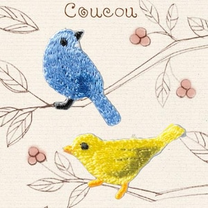 Può includere: Due toppe ricamate a forma di uccellino, una blu e una gialla, con occhi e becchi neri. Le toppe sono mostrate su uno sfondo chiaro con un disegno a linee marroni di rami, foglie e bacche, con la parola Coucou scritta sopra.
