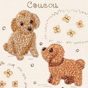 Pode incluir: Dois emblemas bordados apresentando cães de pelo encaracolado castanho-claro sobre um fundo de tecido creme. A palavra "Coucou" está bordada em fio castanho acima dos cães, acompanhada por pequenos motivos florais e contornos circulares dispersos.