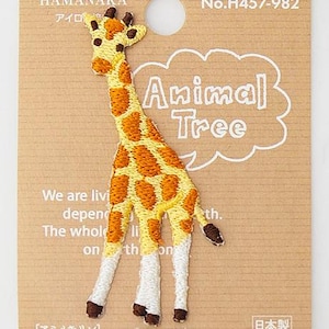 Puede incluir: Un parche bordado de jirafa con manchas amarillas y marrones sobre un soporte de cartón marrón claro. El empaque presenta el texto Animal Tree, Hamanaka, No.H457-982 y texto en inglés sobre la vida en la tierra. La jirafa tiene patas blancas.