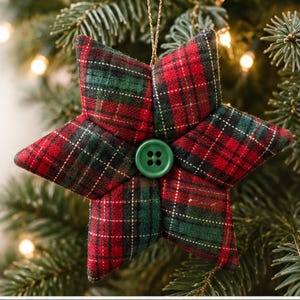 May include: A six-pointed star ornament made of fabric with a red, green, and white plaid pattern. The center of the star features a solid green four-hole button. A thin gold string is attached to the top point for hanging the decoration.