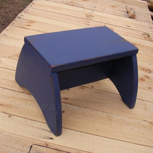 Puede incluir: Un taburete de madera azul marino con una parte superior rectangular y patas curvas. El taburete tiene un acabado envejecido, con desgaste visible en los bordes. El taburete est&aacute; dise&ntilde;ado para uso dom&eacute;stico.