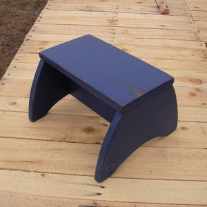 Puede incluir: Un peque&ntilde;o taburete de madera azul marino con un acabado ligeramente envejecido. El taburete tiene una parte superior rectangular y patas curvas. La veta de la madera es visible. Mide unos 40 cm de largo.