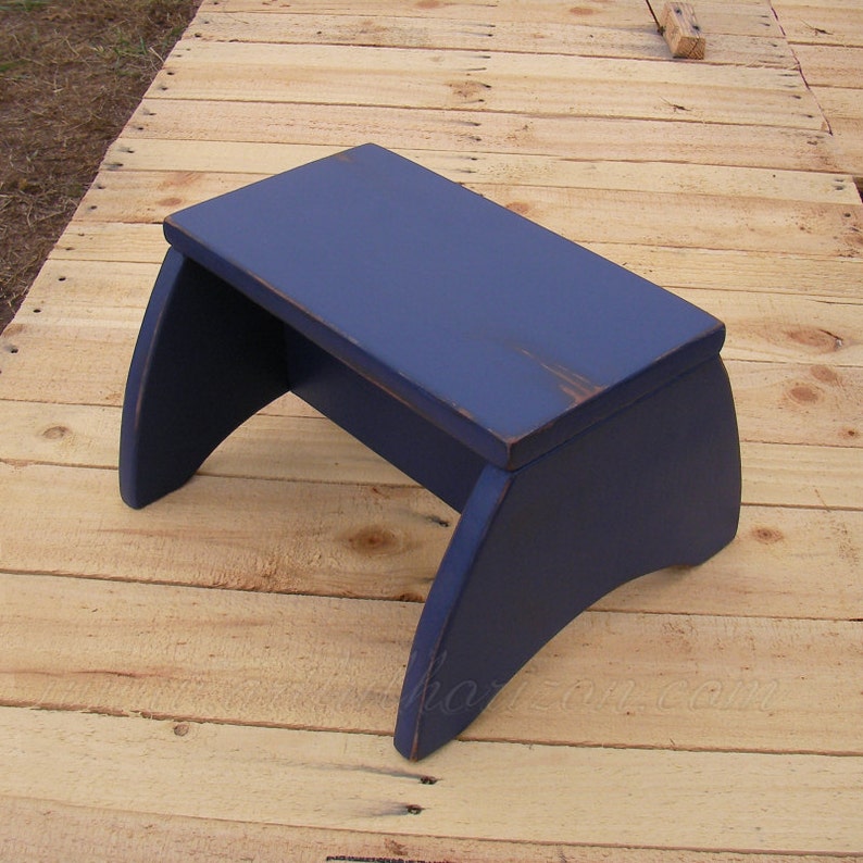 Puede incluir: Un peque&ntilde;o taburete de madera azul marino con un acabado ligeramente envejecido. El taburete tiene una parte superior rectangular y patas curvas. La veta de la madera es visible. Mide unos 40 cm de largo.