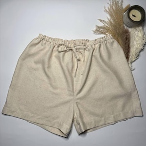 May include: Beige shorts featuring an elasticized waistband with a matching fabric drawstring tie. The garment is crafted from a solid, light-colored textile with a visible weave, showcasing a simple, clean design with a relaxed fit and hemmed edges.