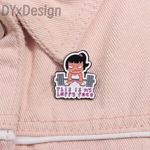 May include: A white and black enamel pin featuring a cartoon girl lifting weights with the text "This is my happy face." The pin is set against a pink background.