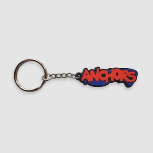 May include: A keychain featuring a metal split ring attached to a short chain and a rubberized charm. The charm displays the word ANCHORS in bold red capital letters with a thick blue outline, set against a dark blue, irregular shaped background.