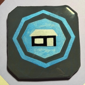 May include: A square, dark gray coaster with a light blue and black house design. The house is white with a black door and window. The coaster has a glossy finish and a geometric shape.