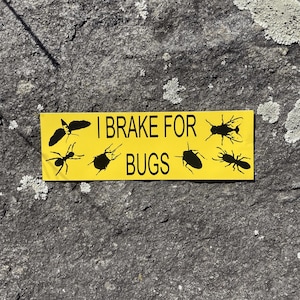 May include: A rectangular yellow bumper sticker featuring the black text I BRAKE FOR BUGS in the center. The design includes six black silhouettes of various insects, including a moth, an ant, a beetle, and a fly, arranged around the text.