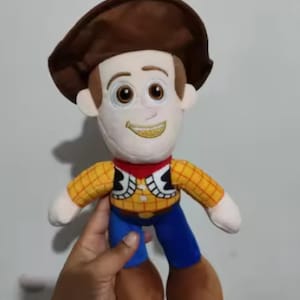 May include: A plush toy of a cartoon cowboy character. The toy has a brown hat, a white face with embroidered features, a yellow plaid shirt, blue pants, and brown boots. The character has a red neckerchief and a yellow embroidered smile.