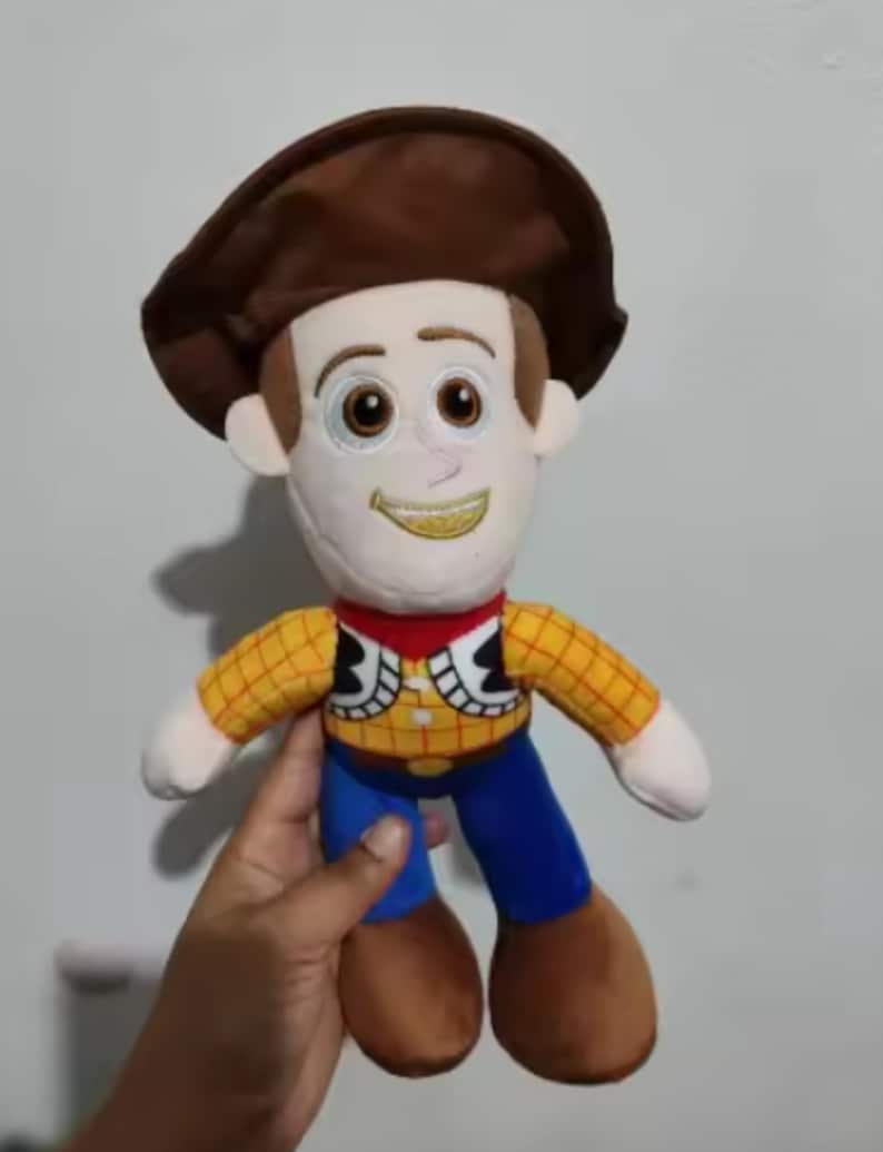 May include: A plush toy of a cartoon cowboy character. The toy has a brown hat, a white face with embroidered features, a yellow plaid shirt, blue pants, and brown boots. The character has a red neckerchief and a yellow embroidered smile.