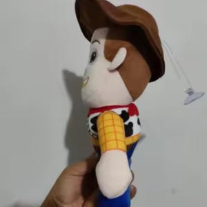 May include: A plush toy of the character Woody from the animated film Toy Story. The doll features a brown cowboy hat, a yellow plaid shirt, a red bandana, and blue pants. A small suction cup is attached.