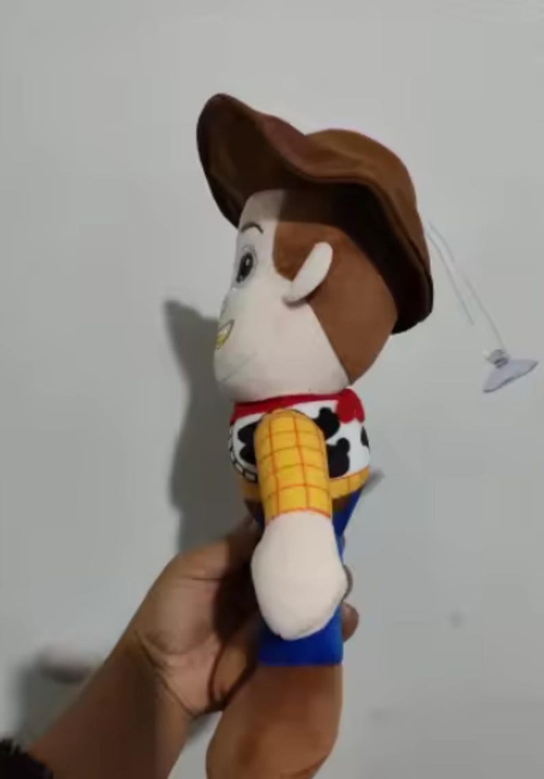 May include: A plush toy of the character Woody from the animated film Toy Story. The doll features a brown cowboy hat, a yellow plaid shirt, a red bandana, and blue pants. A small suction cup is attached.