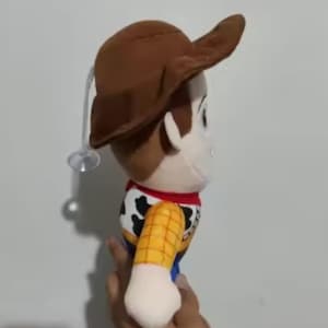 May include: A plush toy of the character Woody from the movie Toy Story. The doll has a brown cowboy hat, a yellow shirt with a cow print vest, blue pants, and a red neckerchief. The toy is held in a hand.