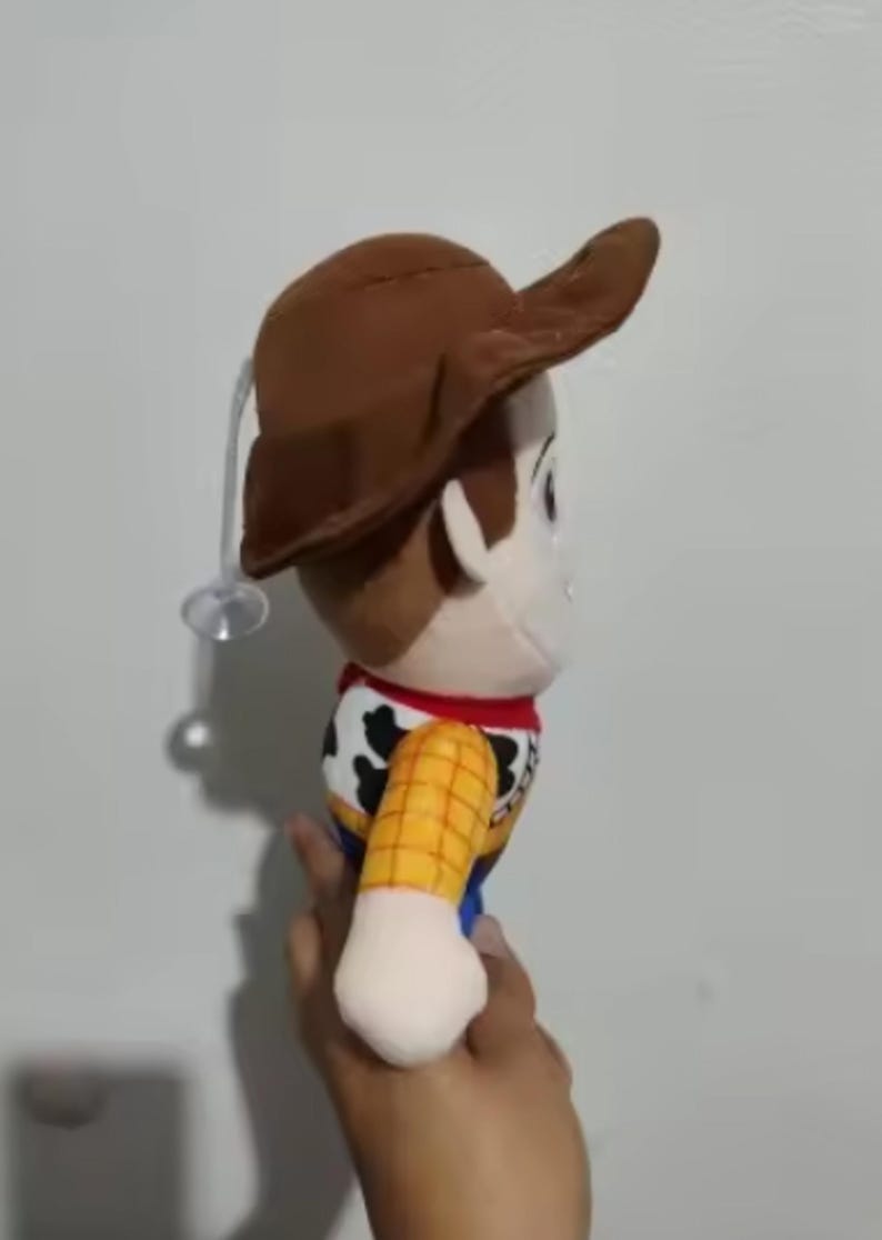 May include: A plush toy of the character Woody from the movie Toy Story. The doll has a brown cowboy hat, a yellow shirt with a cow print vest, blue pants, and a red neckerchief. The toy is held in a hand.