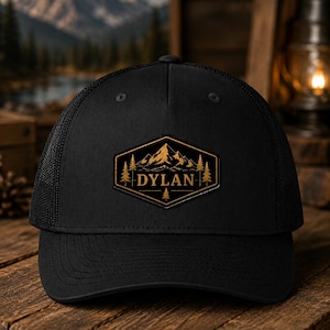 May include: Black trucker cap with a mesh back and a curved peak. The front displays a gold and black mountain and tree design with the name "DYLAN" in gold lettering. The cap is on a wooden surface.