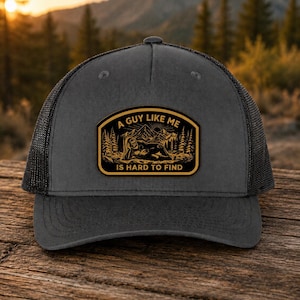 May include: A grey trucker cap with a black mesh back. The cap has a rectangular patch with the text "A GUY LIKE ME IS HARD TO FIND" and a graphic of a person relaxing in front of a mountain scene.