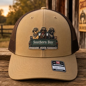 May include: A tan trucker hat with a brown mesh back. The front features a patch with three dogs in a truck bed, the text "Southern Boy" and "USA". The hat has an adjustable strap and a Richardson 112FP Trucker tag.