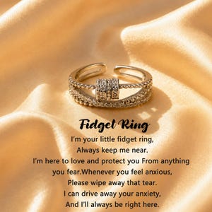 May include: A silver-toned metal fidget ring featuring a textured band with three small, rotating cylindrical beads. The ring is accompanied by the printed text: Fidget Ring, I'm your little fidget ring, Always keep me near. I'm here to love and protect you From anything you fear. Whenever you feel anxious, Please wipe away that tear. I can drive away your anxiety, And I'll always be right here.