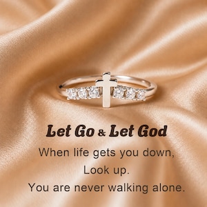 May include: A silver-toned ring featuring a central cross emblem flanked by three small, clear, round-cut stones on each side. The text "Let Go & Let God. When life gets you down, Look up. You are never walking alone." is printed below the ring.