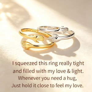 May include: Two open-ended rings, one gold-toned and one silver-toned, shaped like two hands embracing. The gold ring is engraved with the text 'Love you forever' and the silver ring with 'Love you'. Below, text reads: 'I squeezed this ring really tight and filled with my love & light. Whenever you need a hug, Just hold it close to feel my love.'