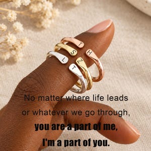 May include: Three open-ended rings in rose gold, yellow gold, and silver tones, each featuring a single stamped letter. The rings are stacked on a finger. Text overlay reads: No matter where life leads or whatever we go through, you are a part of me, I'm a part of you.