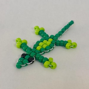May include: A beaded lizard figurine in shades of green and yellow-green. The lizard's body is made of green beads, with lighter green beads on the legs and head. Black beads form the eyes. The beads are strung together with white string.