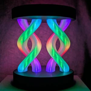 May include: A decorative lamp featuring two translucent, twisted, double-helix structures illuminated with vibrant rainbow gradients of pink, green, and blue light, set between two solid black circular bases at the top and bottom.