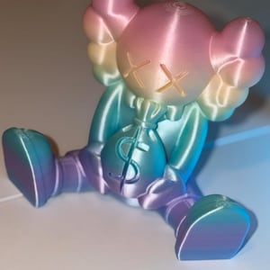 May include: A colorful figurine in a seated position, holding a money bag. The figure has a rounded head with crossed-out eyes and large, rounded ears. The color gradient shifts from pink to blue and purple. The money bag has a dollar sign.