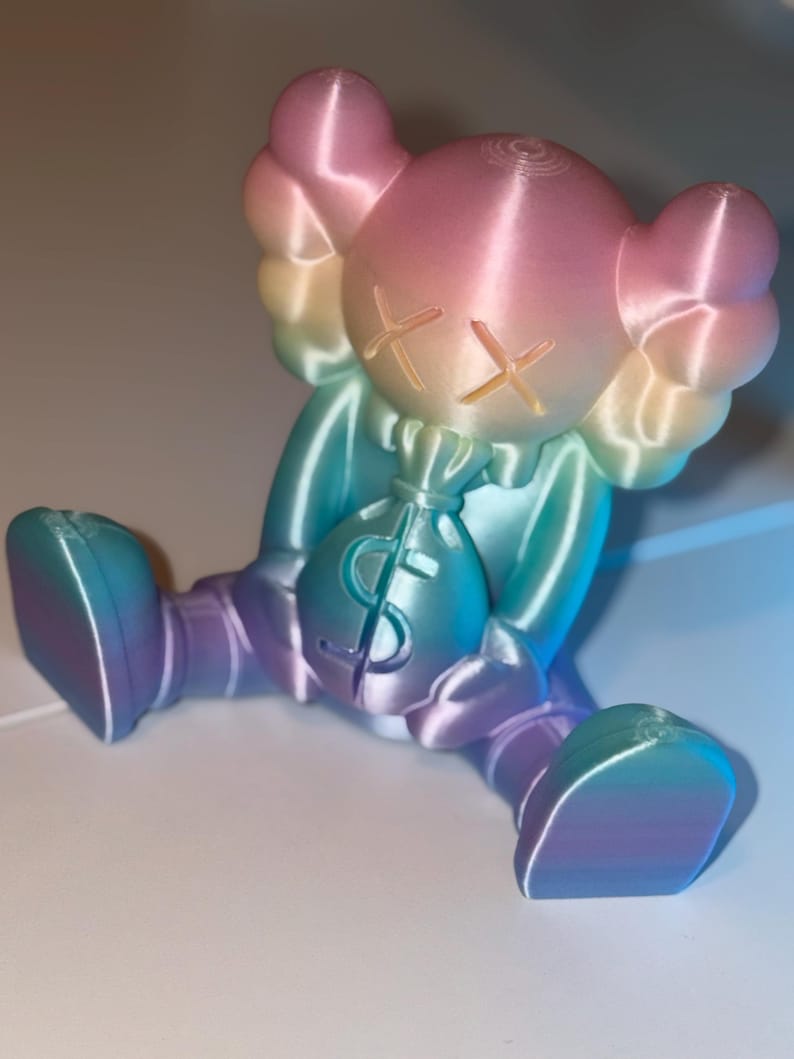 May include: A colorful figurine in a seated position, holding a money bag. The figure has a rounded head with crossed-out eyes and large, rounded ears. The color gradient shifts from pink to blue and purple. The money bag has a dollar sign.